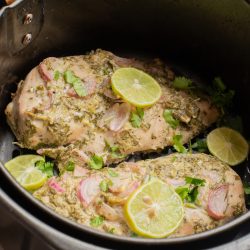 Two seasoned chicken breasts, inspired by Marinated Yogurt Herb Chicken, are topped with lime slices and herbs, then cooked to perfection in a black slow cooker.
