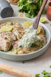 A wooden spoon holds creamy herb sauce over Air Fryer Cilantro Lime Chicken—marinated breasts garnished with lime slices and cilantro in a shallow bowl.