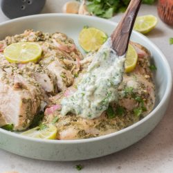 A wooden spoon holds creamy herb sauce over Air Fryer Cilantro Lime Chicken—marinated breasts garnished with lime slices and cilantro in a shallow bowl.