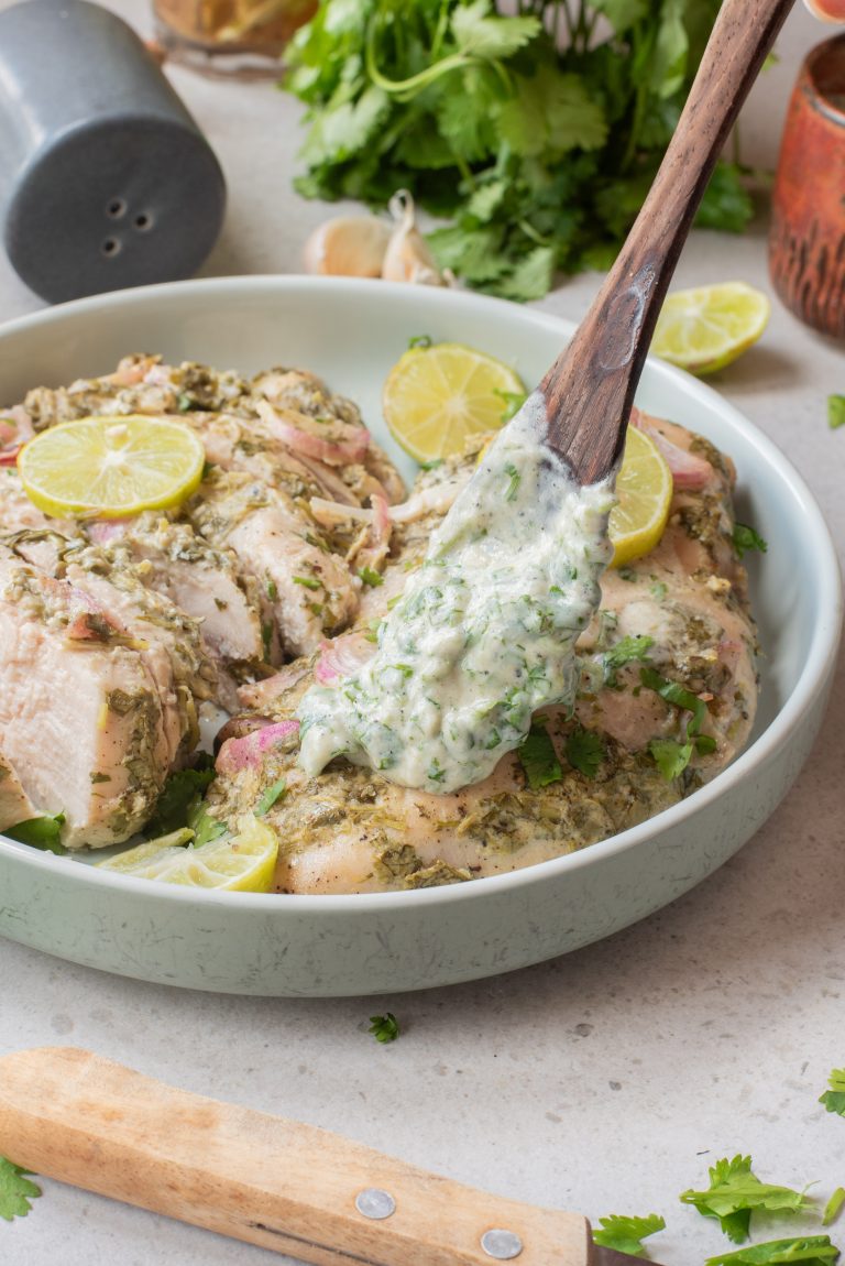 A wooden spoon holds creamy herb sauce over Air Fryer Cilantro Lime Chicken—marinated breasts garnished with lime slices and cilantro in a shallow bowl.