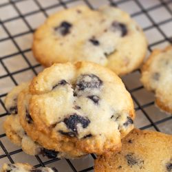 A close-up of several Dried Blueberry Lemon Cookies cooling on a black wire rack, their golden edges and fruity aroma inviting you to enjoy a fresh-baked treat.