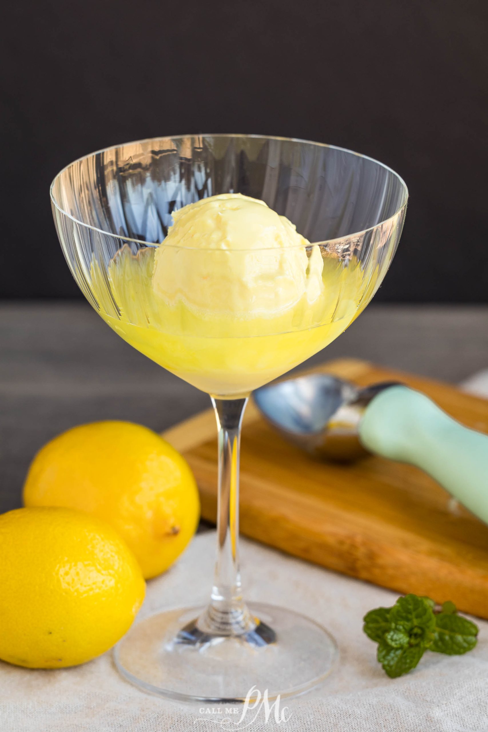 A glass with a scoop of lemon sorbet, perfect for creating a refreshing limoncello float, with two whole lemons, a mint sprig, and an ice cream scoop on a wooden board in the background.