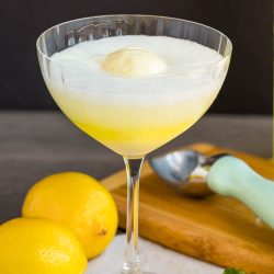 A coupe glass filled with a yellow fizzy limoncello float topped with vanilla ice cream, surrounded by two lemons, an ice cream scoop, and a fresh mint sprig.