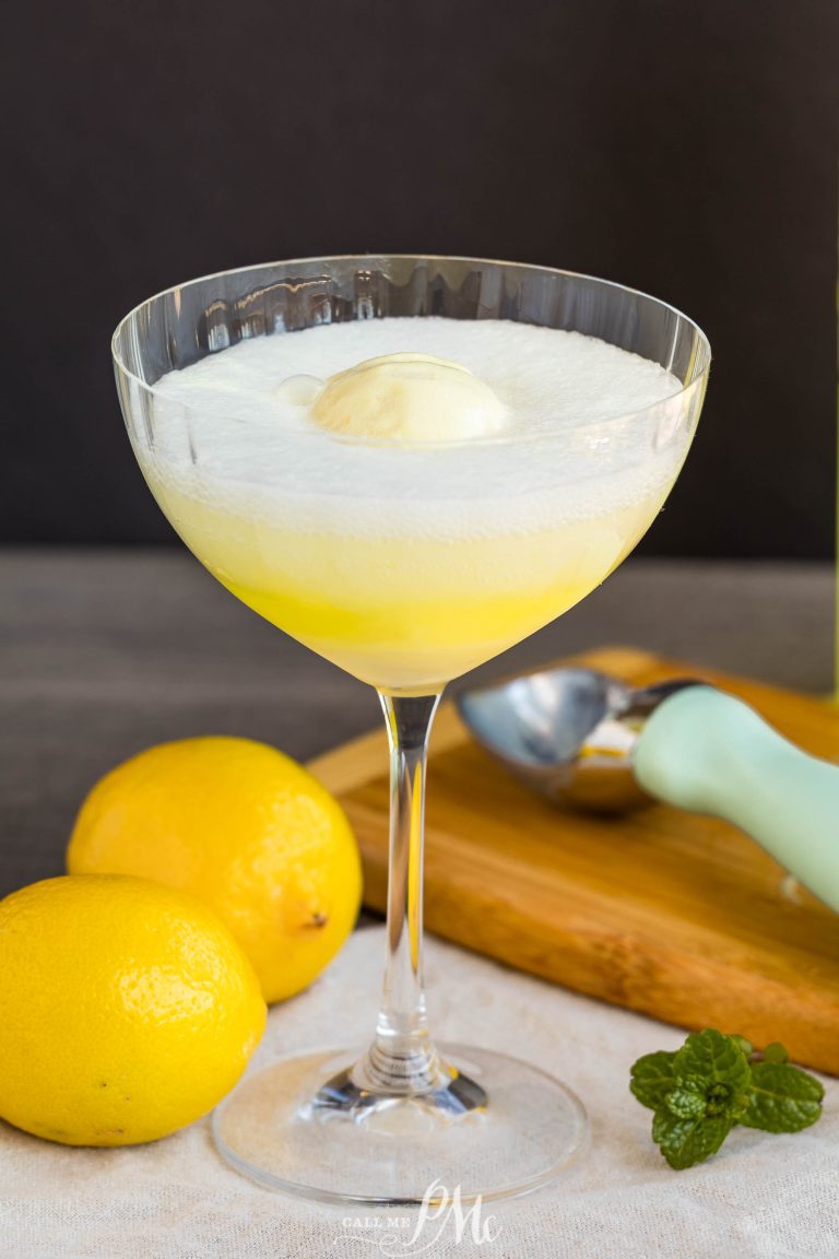 A coupe glass filled with a yellow fizzy limoncello float topped with vanilla ice cream, surrounded by two lemons, an ice cream scoop, and a fresh mint sprig.