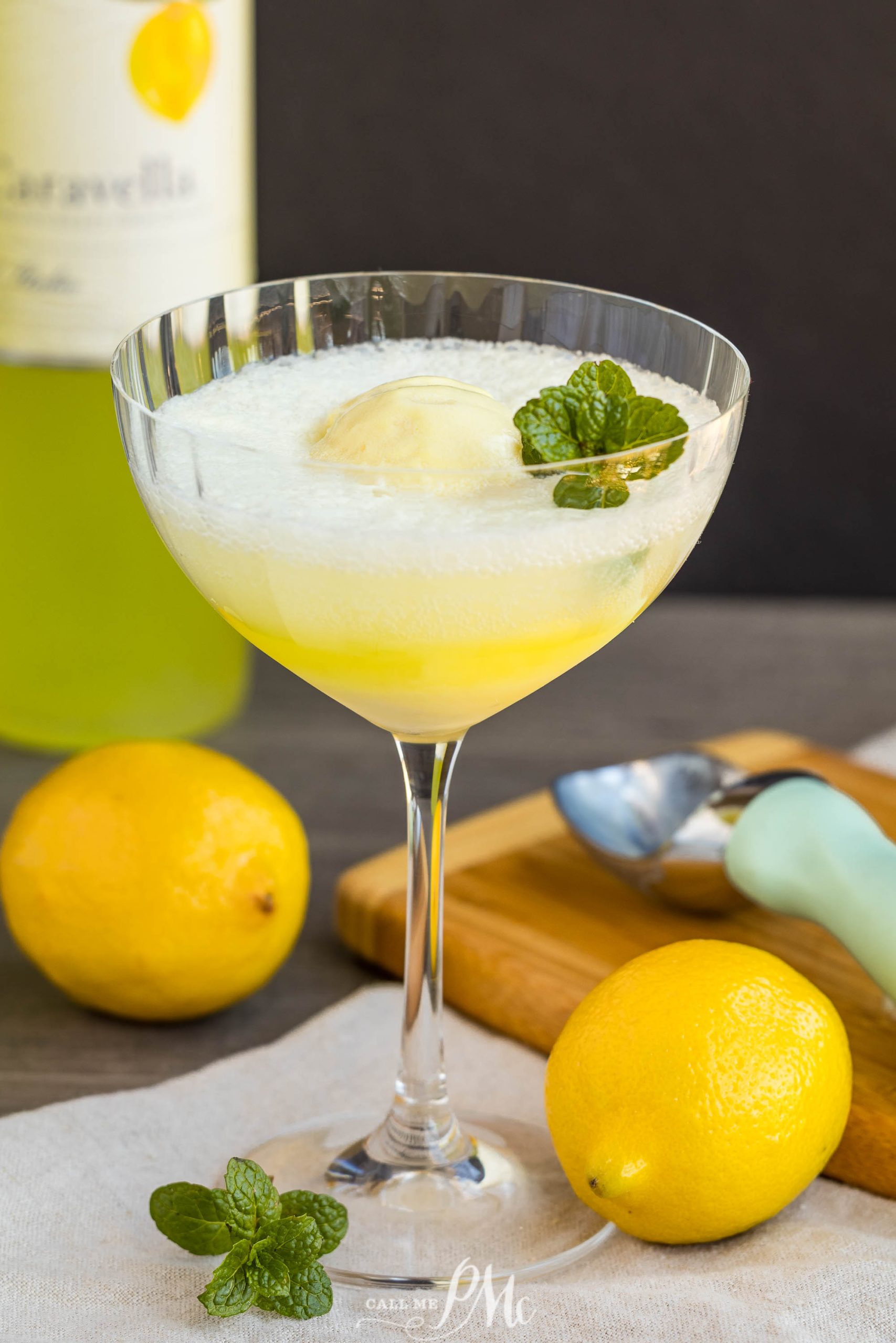 A cocktail glass filled with a yellow liquid, topped with a mint leaf, sits on a table surrounded by lemons, a bottle, and an ice cream scoop.