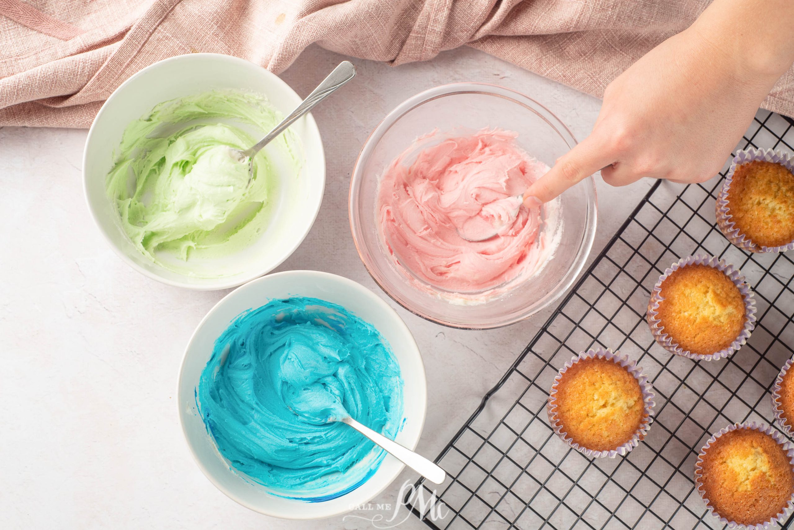Three bowls of pastel-colored frosting (green, pink, blue) with spoons, a hand touching pink frosting, and plain cupcakes cooling on a wire rack.