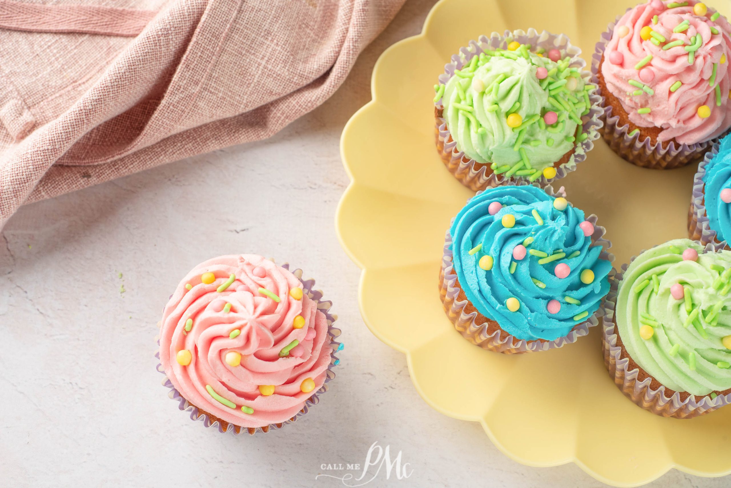 A yellow plate with Easter vanilla cupcakes decorated in pink, blue, and green frosting with sprinkles sits on a light surface next to a single pink cupcake and a pink cloth.