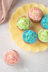 Six cupcakes with pink, green, and blue frosting and colorful sprinkles are arranged on a yellow plate, with one green and pink cupcake placed next to the plate.