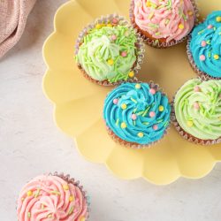 Six cupcakes with pink, green, and blue frosting and colorful sprinkles are arranged on a yellow plate, with one green and pink cupcake placed next to the plate.