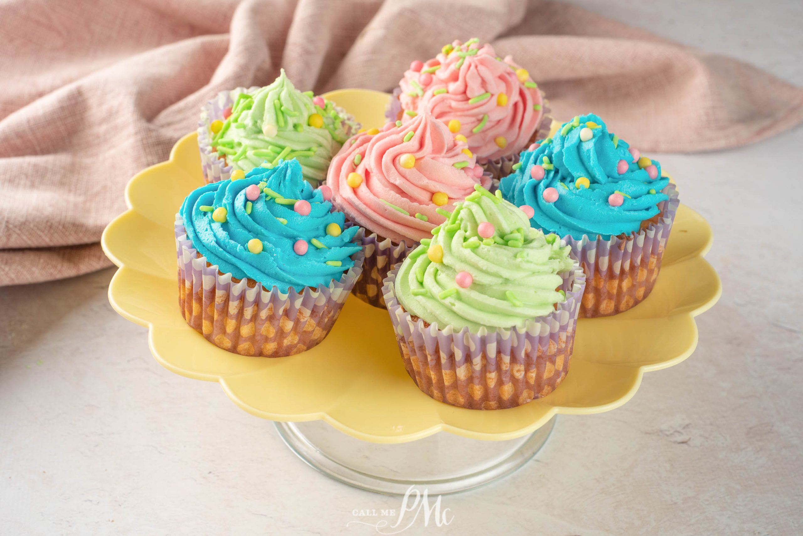 Six Easter vanilla cupcakes with pink, blue, and green frosting and colorful sprinkles are arranged on a yellow scalloped cake stand.