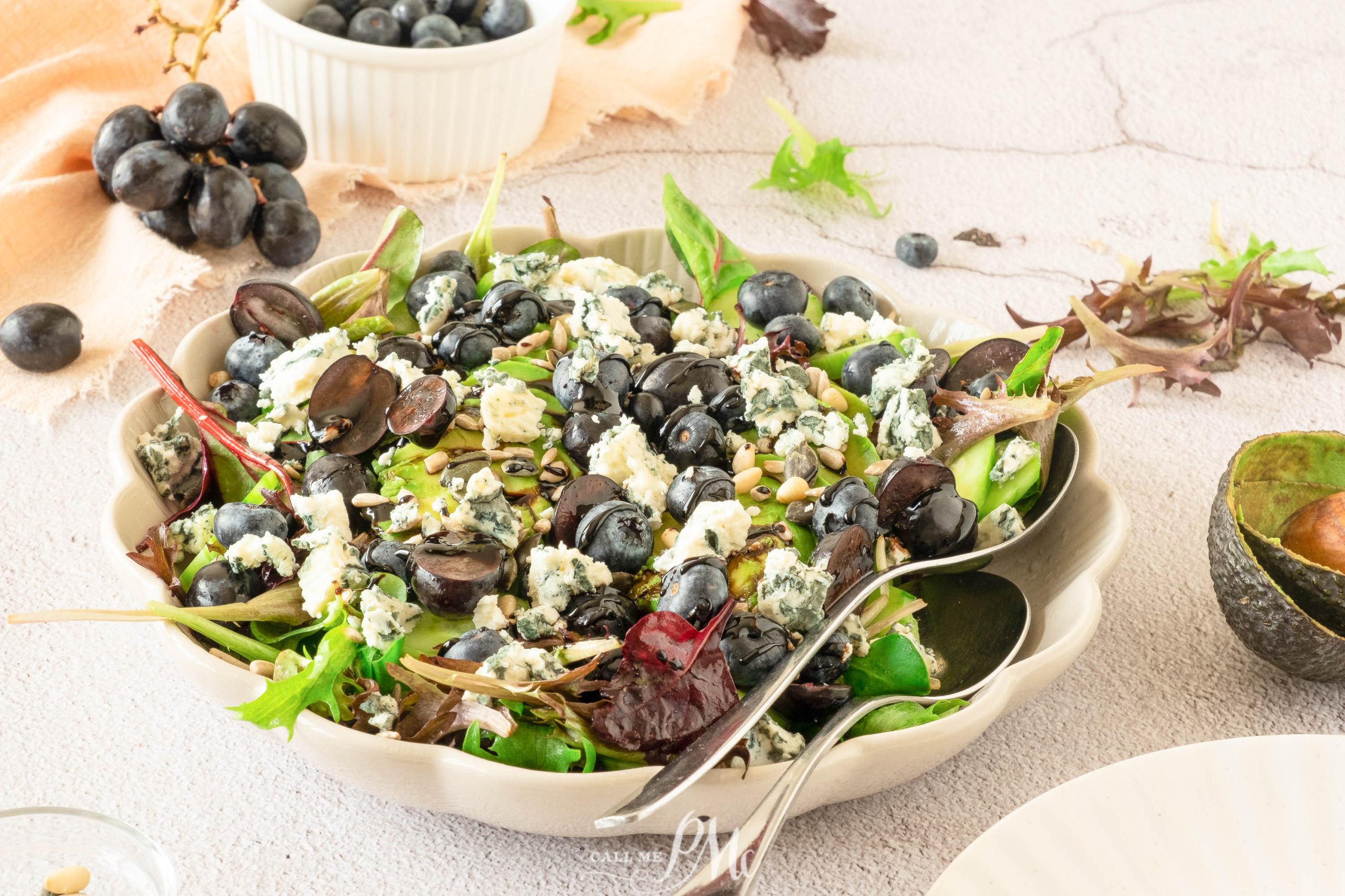 A vibrant Avocado Blueberry Arugula Salad featuring mixed greens, black grapes, blueberries, crumbled blue cheese, and seeds in a bowl, surrounded by extra grapes and an avocado half.