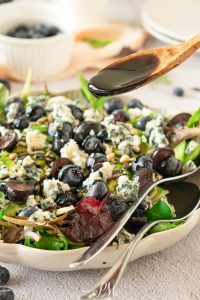 A close-up of a salad with mixed greens, blueberries, blue cheese, and pine nuts, as a wooden spoon drizzles dark balsamic dressing over the top.