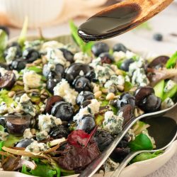 A close-up of a salad with mixed greens, blueberries, blue cheese, and pine nuts, as a wooden spoon drizzles dark balsamic dressing over the top.