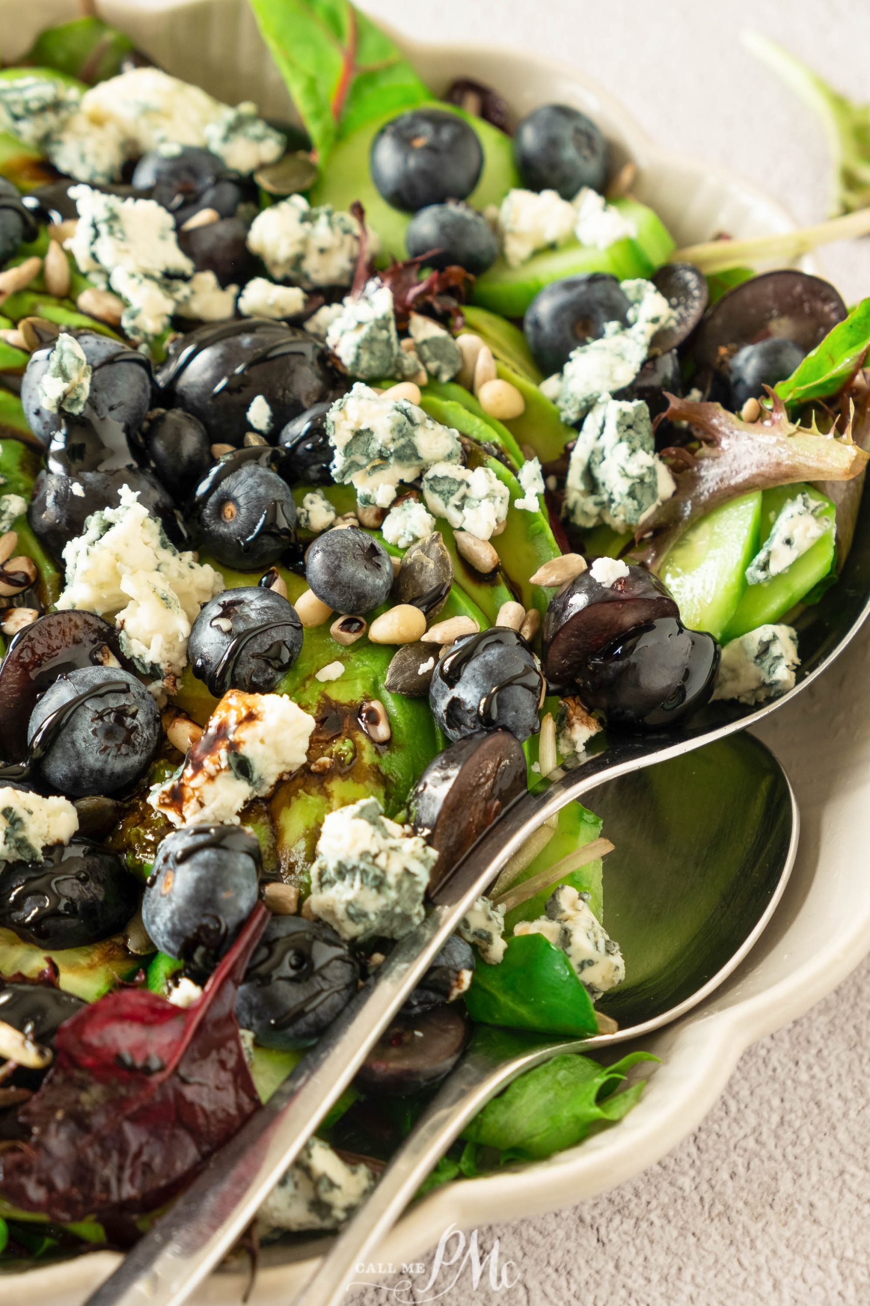 Salad with mixed greens, blueberries, crumbled blue cheese, pine nuts, and a drizzle of dressing in a bowl with serving utensils.