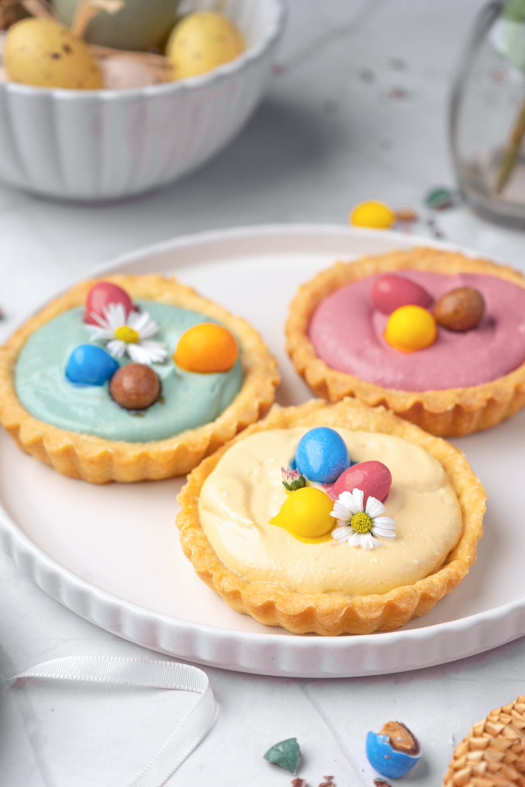 Three desserts with pastel-colored fillings, topped with candy eggs and small flowers, are arranged on a white plate. A bowl of eggs is visible in the background.