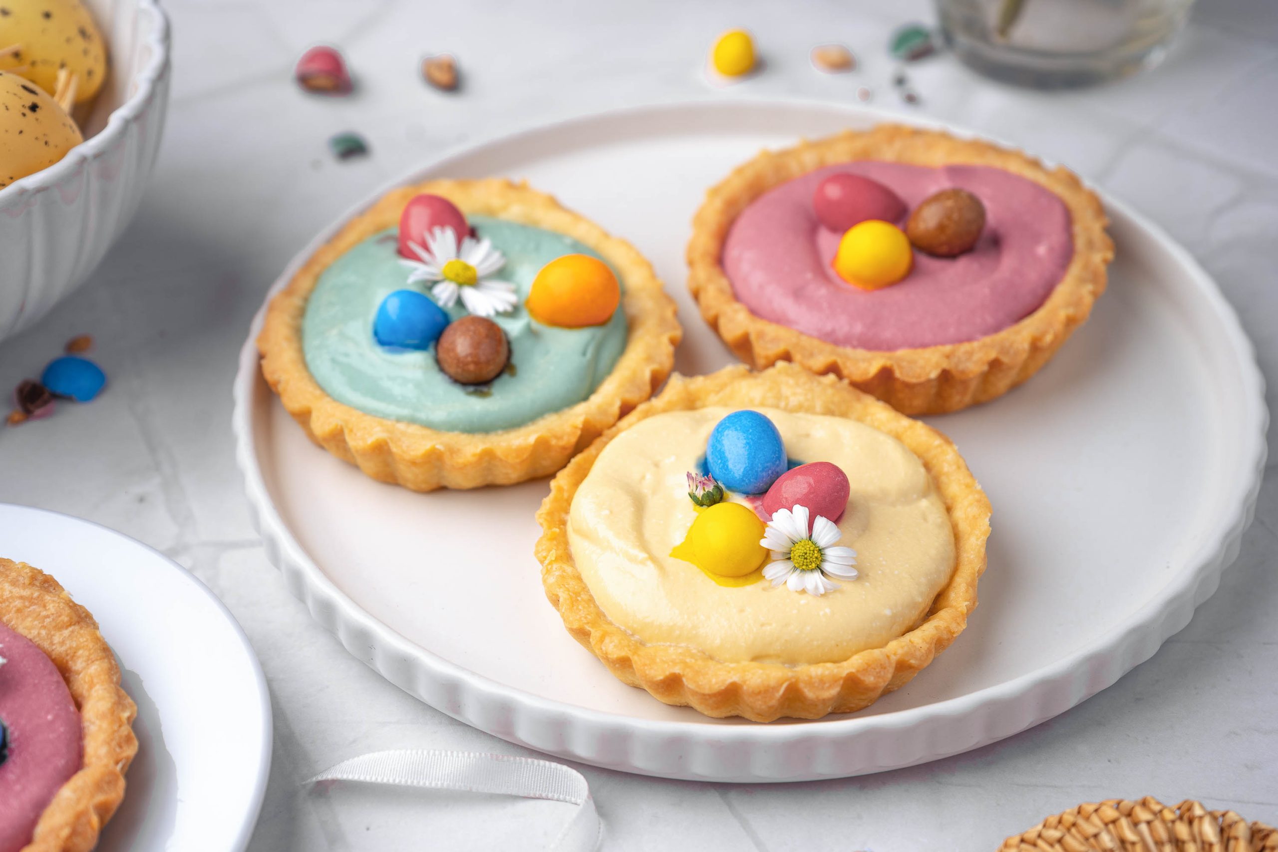 Three pastel-colored Springtime Cheesecake Tartlets on a white plate, topped with small candy eggs and white edible flowers, with scattered candies in the background.