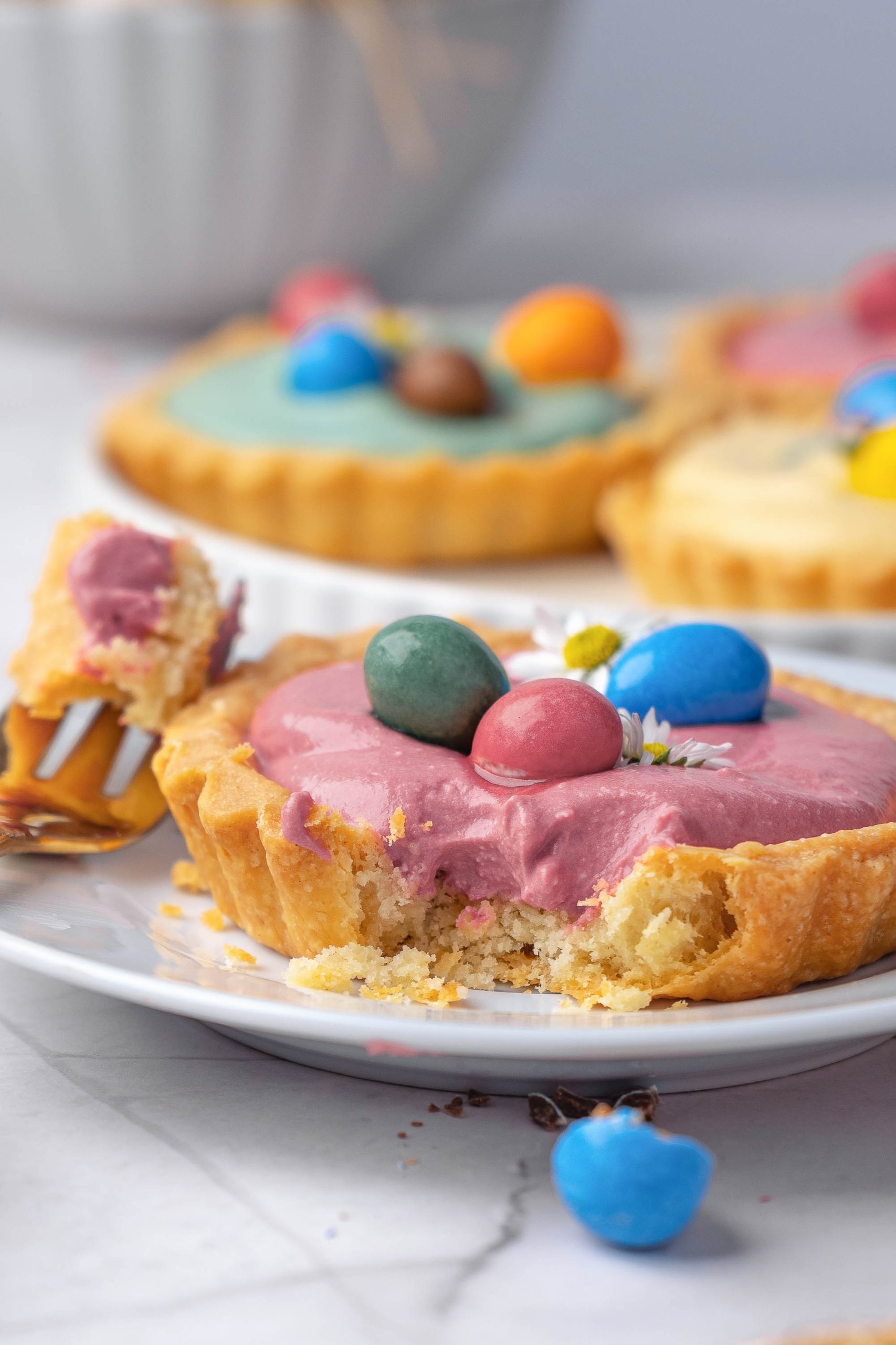 Desserts with pink filling and colorful candy eggs on top sits on a plate with a bite taken out and a fork holding a piece; more tartlets are in the background.