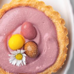 A Springtime Cheesecake Tartlet with pink cream filling, topped with three colorful candy eggs and a small white daisy flower, served on a white plate.