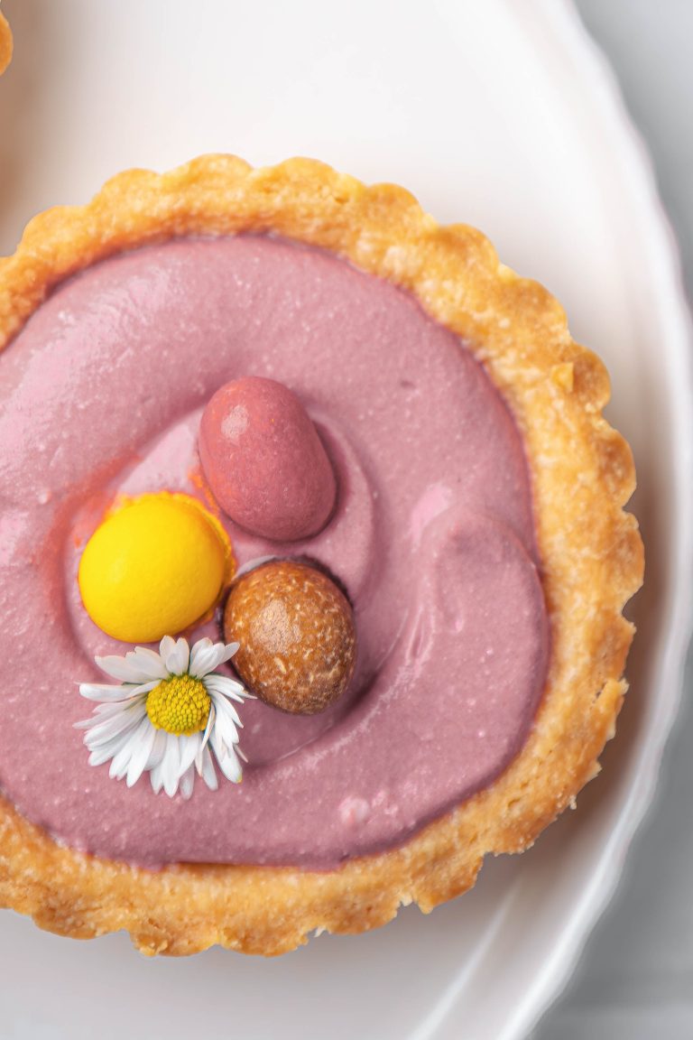 A Springtime Cheesecake Tartlet with pink cream filling, topped with three colorful candy eggs and a small white daisy flower, served on a white plate.