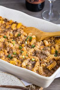 A white baking dish filled with cooked rotini pasta mixed with ground beef and sauce, topped with chopped green onions. A wooden spoon rests in the dish.