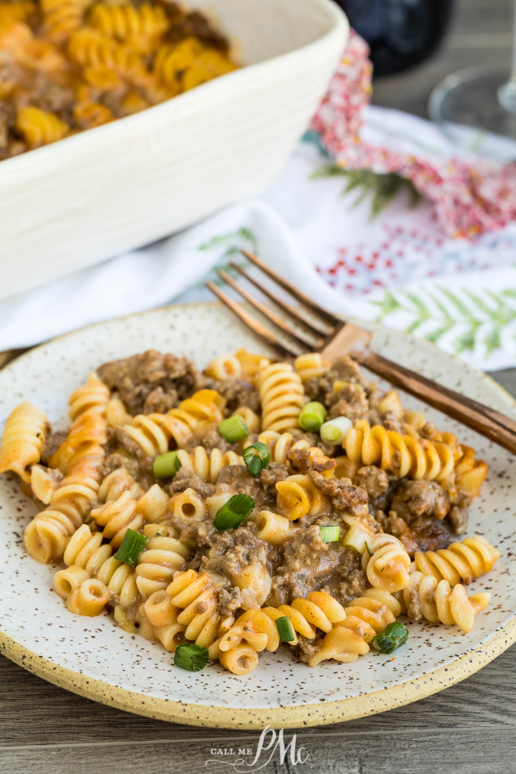 A plate of rotini pasta casserole with ground beef, sauce, and chopped green onions, served with a fork on a speckled plate.