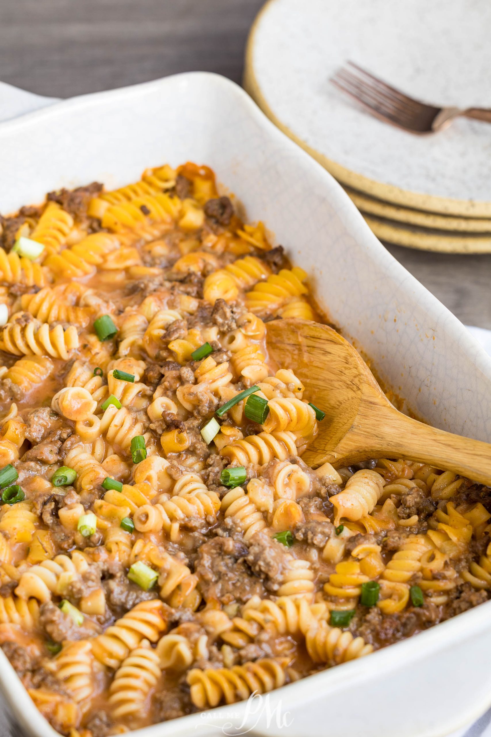 A baked pasta casserole with rotini, ground meat, cheese, and chopped green onions in a white dish with a wooden spoon. Plates and a fork are in the background.