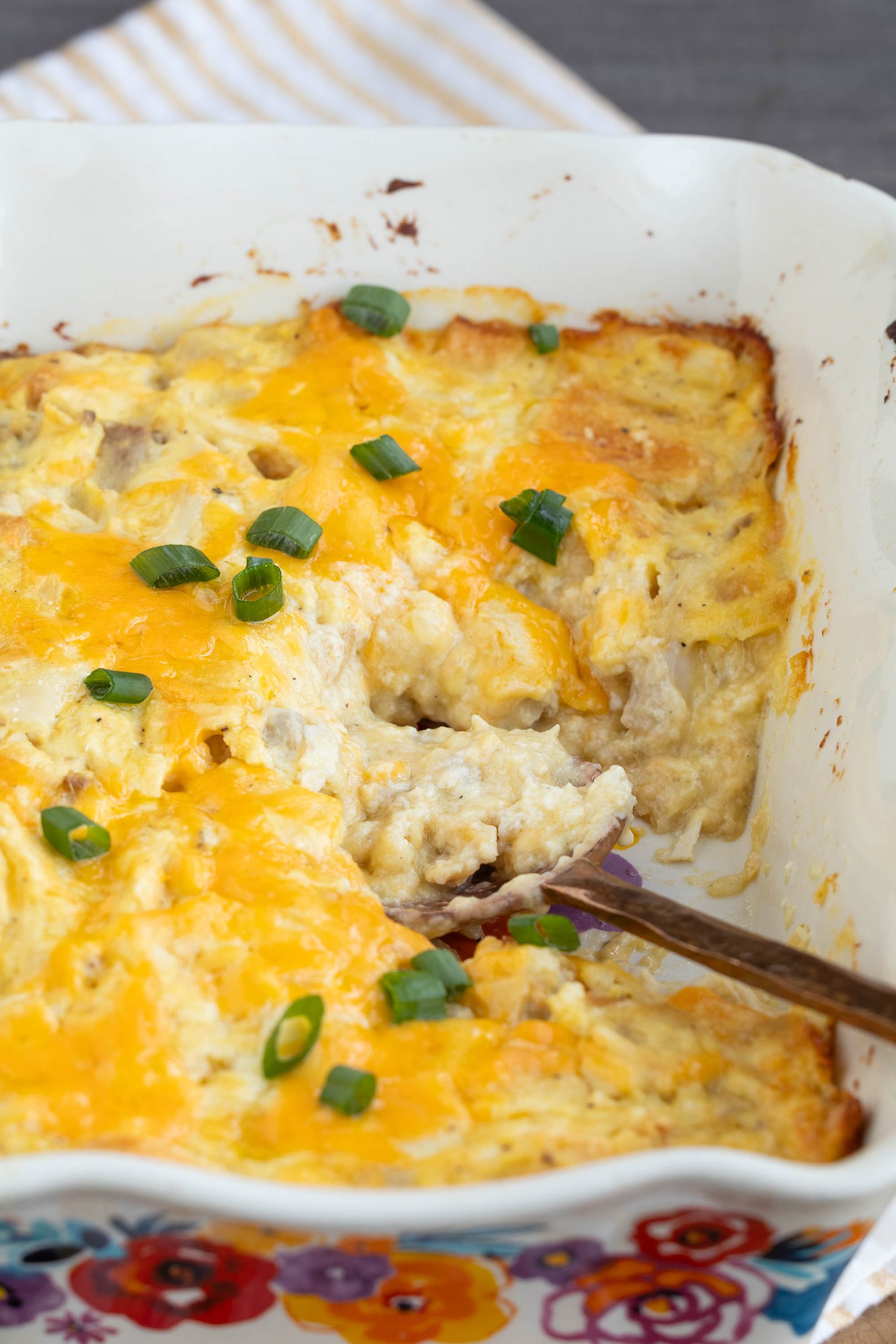A baked dish topped with melted cheddar cheese and green onions, with a serving spoon scooping out a portion from a white baking dish.