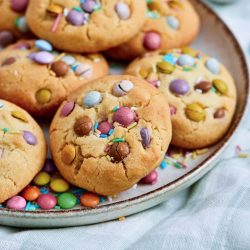 A plate of round Easter M&M Cookies topped with colorful candy-coated chocolates and sprinkles, placed on a light fabric surface.