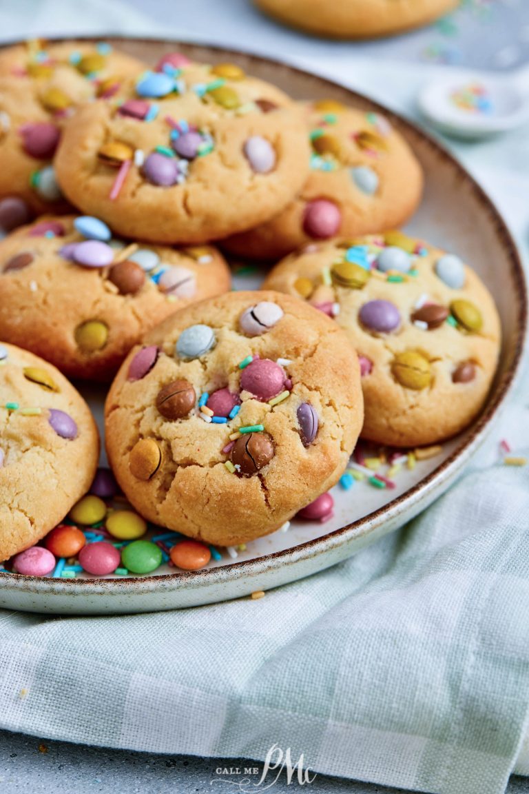 A plate of round Easter M&M Cookies topped with colorful candy-coated chocolates and sprinkles, placed on a light fabric surface.