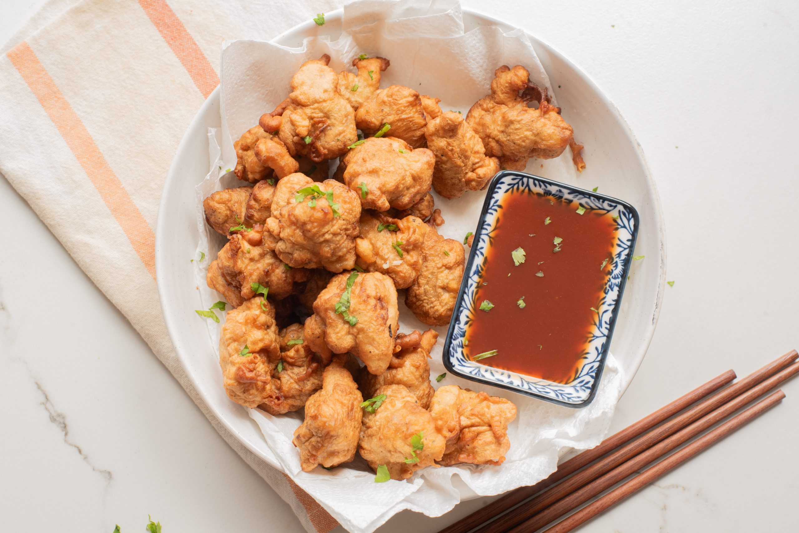A plate of fried battered Chinese chicken bites with chopped herbs, served with a dish of brown dipping sauce; chopsticks are placed beside the plate.