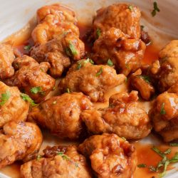 A white bowl filled with battered and fried cauliflower florets coated in a reddish-orange sauce, garnished with chopped herbs.
