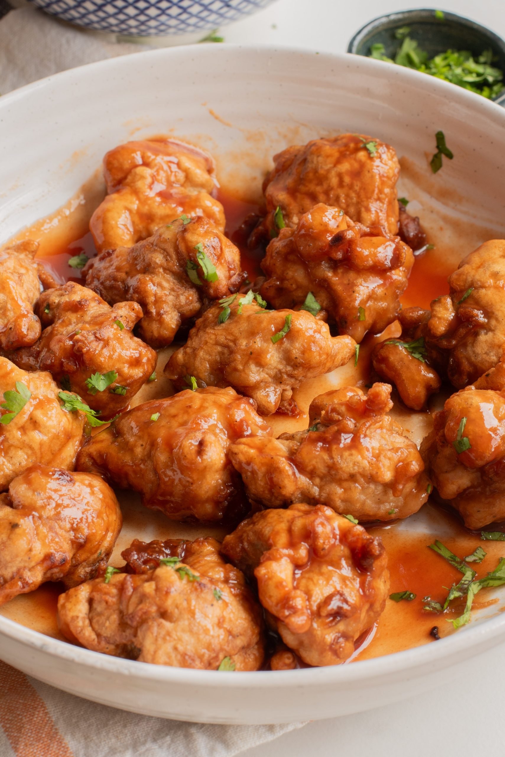 A white bowl filled with battered and fried chicken coated in a reddish-orange sauce, garnished with chopped herbs.