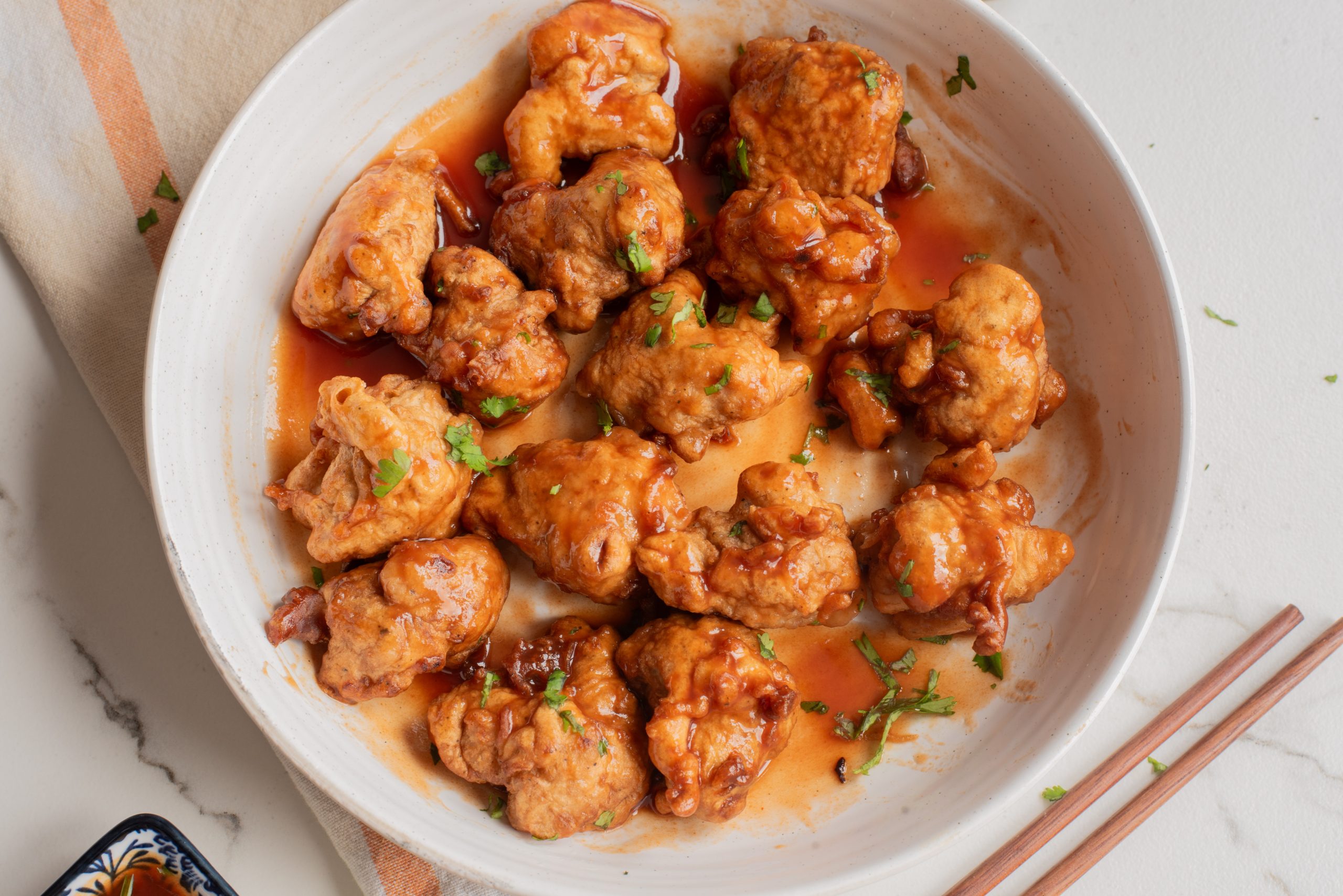 A white bowl filled with battered and fried chicken pieces coated in a reddish-brown sauce, garnished with chopped green herbs. Chopsticks rest nearby on the surface.