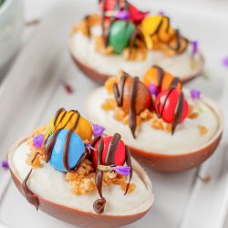Three chocolate egg halves filled with cream, topped with colorful candies, crumbs, edible flowers, and drizzled with chocolate, arranged on a white rectangular plate.
