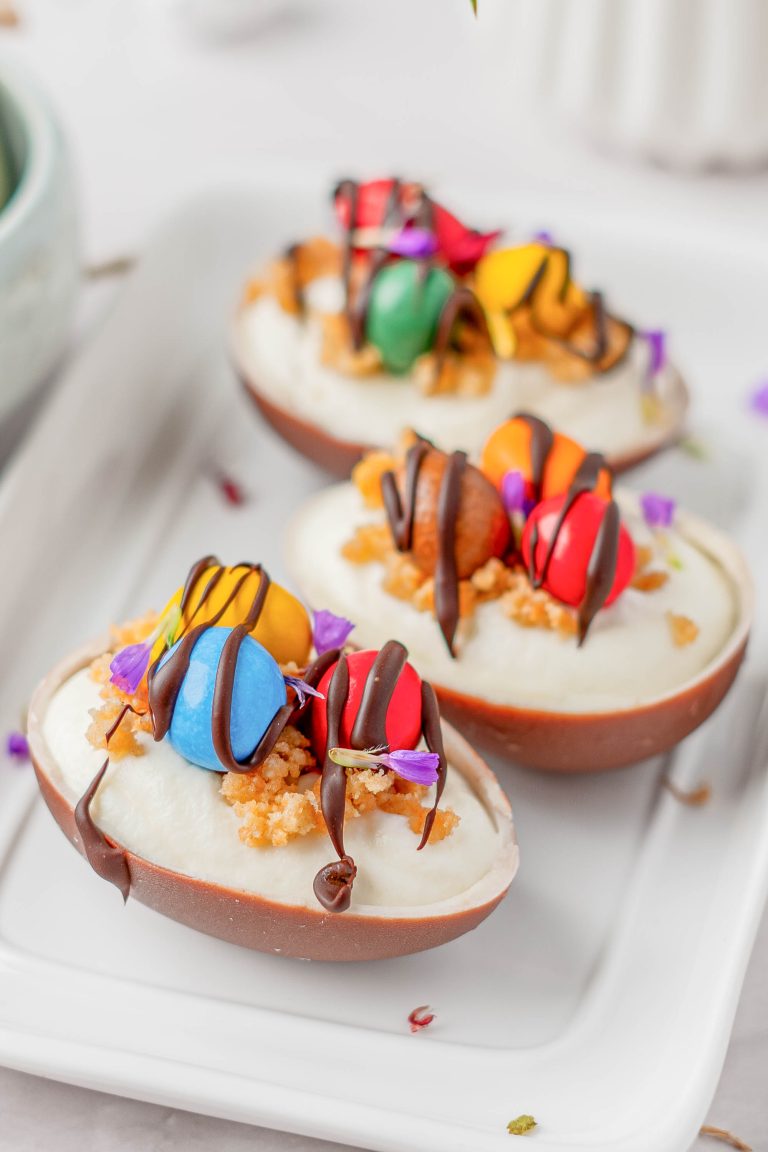 Three chocolate egg halves filled with cream, topped with colorful candies, crumbs, edible flowers, and drizzled with chocolate, arranged on a white rectangular plate.