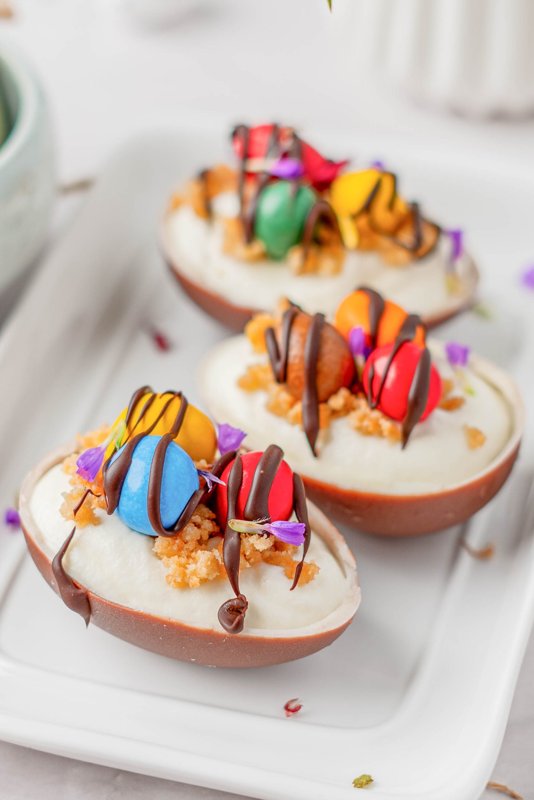 Three Individual Easter Egg Cheesecakes filled with cream, topped with colorful candies, crumbs, edible flowers, and drizzled with chocolate, arranged on a white rectangular plate.