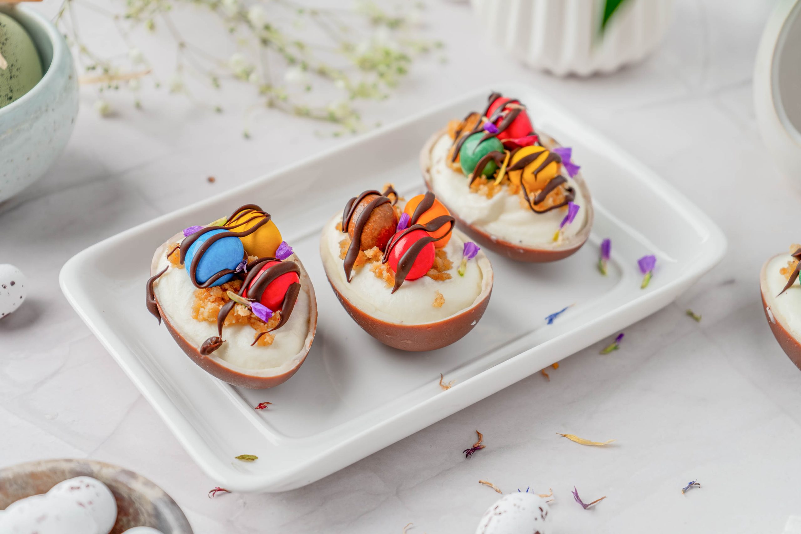 Three chocolate egg halves filled with cream and topped with small colorful macarons, nuts, and flower petals are arranged on a rectangular white plate.