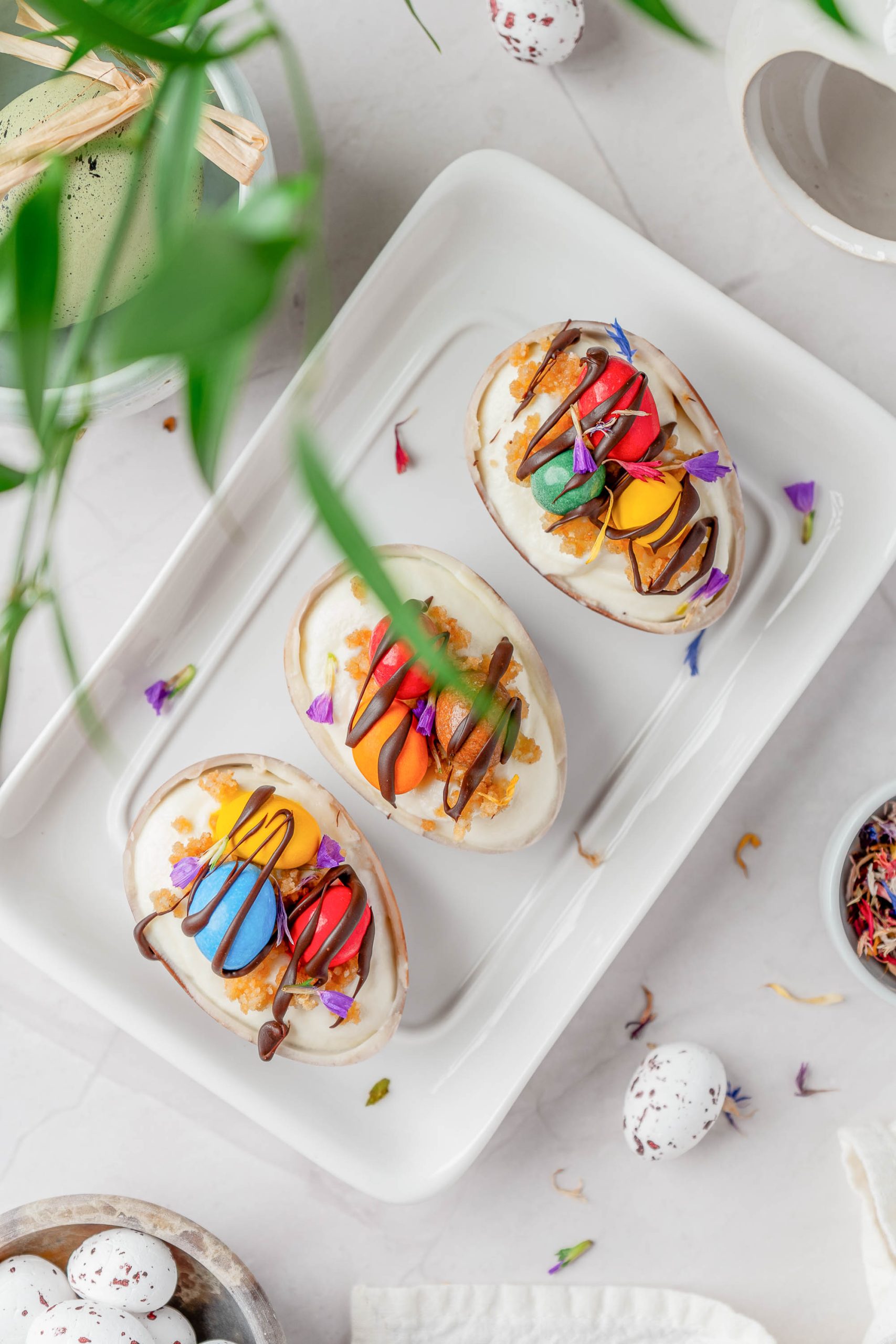 Three chocolate egg halves filled with cream and topped with colorful decorations and drizzled chocolate, arranged on a white rectangular plate.
