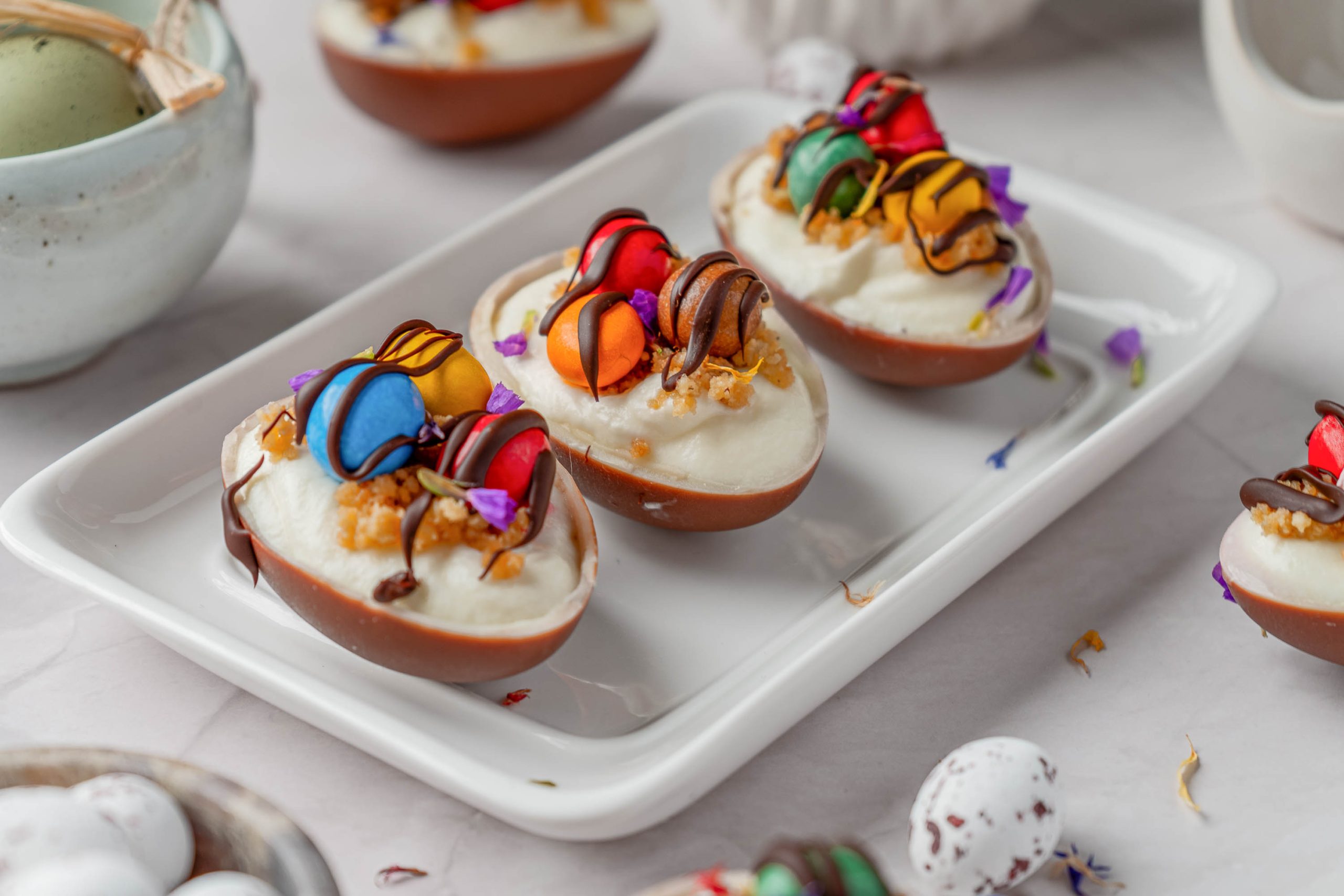 Three chocolate egg halves filled with white cream, topped with colored chocolate hearts, sprinkles, and a drizzle of chocolate, arranged on a rectangular white plate.