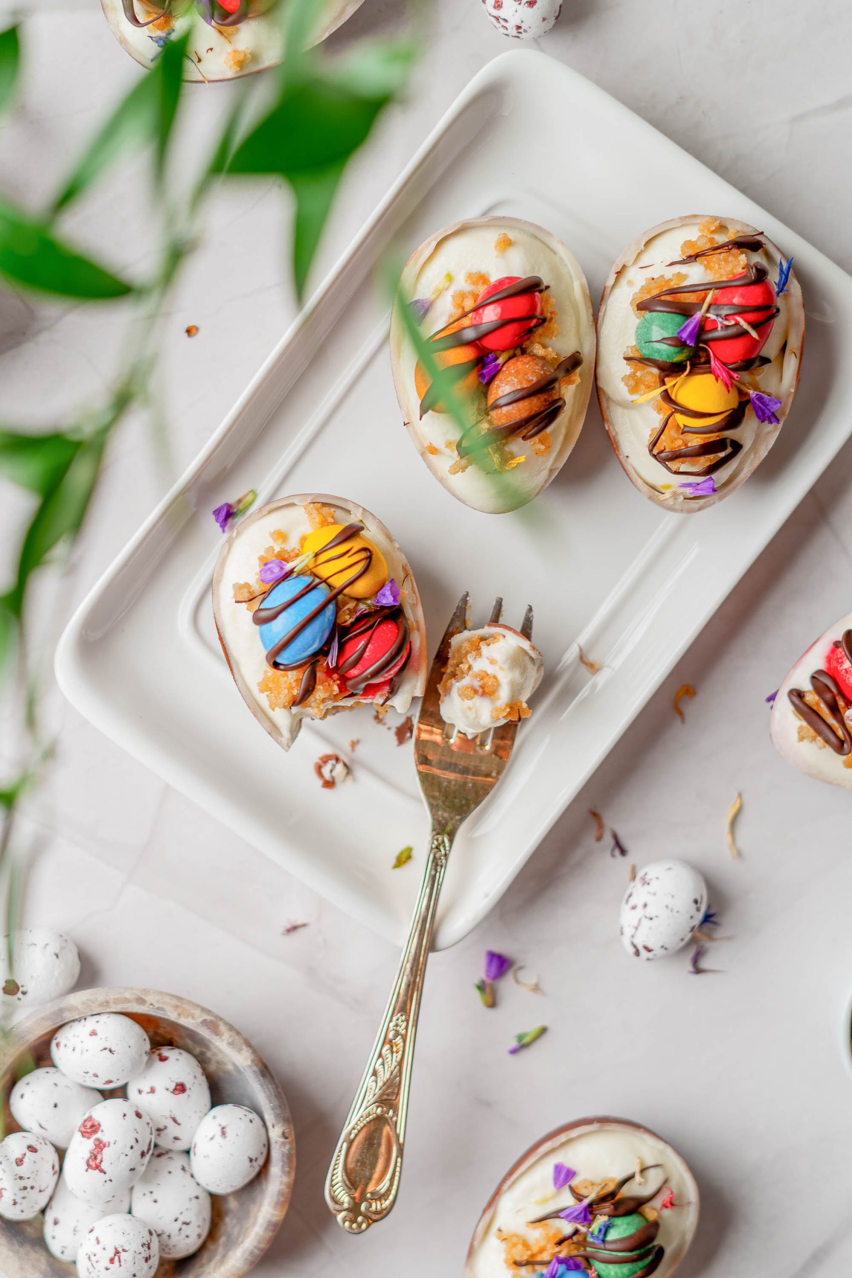 Three decorated Individual Easter Egg Cheesecakes topped with colorful cream, nuts, and edible flowers on a white tray, with a fork and speckled candy eggs nearby.