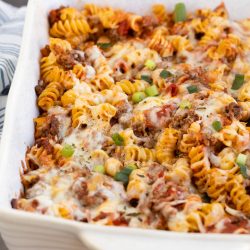 A white baking dish filled with Meat Lovers Pasta Casserole—baked rotini pasta, ground meat, tomato sauce, melted cheese, and garnished with chopped green onions.