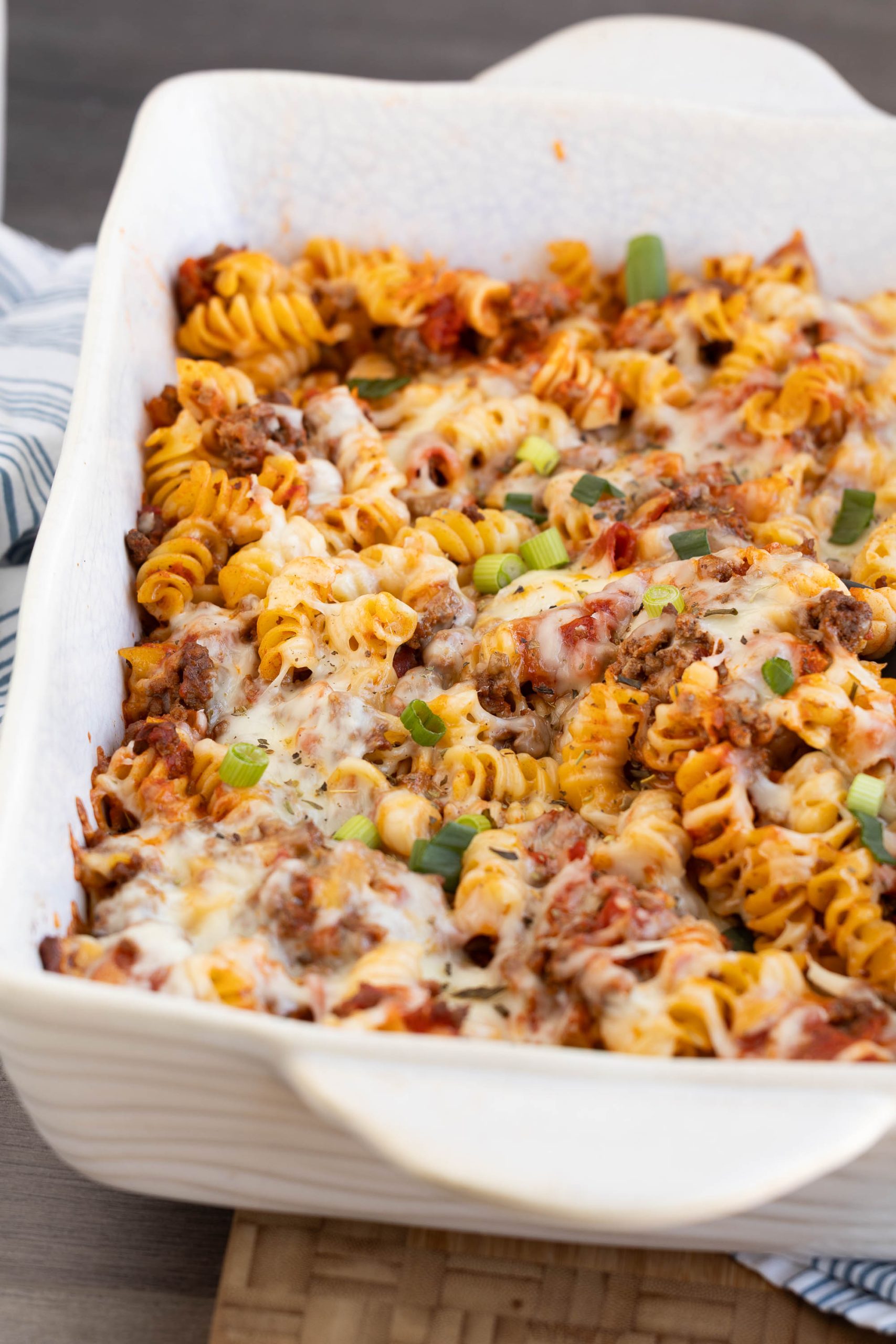 A white baking dish filled with Meat Lovers Pasta Casserole—baked rotini pasta, ground meat, tomato sauce, melted cheese, and garnished with chopped green onions.