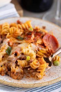 A plate of baked rotini pasta with melted cheese, ground meat, tomato sauce, sliced pepperoni, and chopped green onions, with a fork resting on the plate.