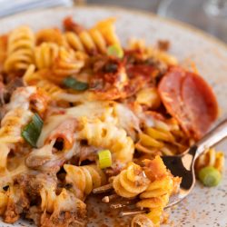 A plate of baked rotini pasta with melted cheese, ground meat, tomato sauce, sliced pepperoni, and chopped green onions, with a fork resting on the plate.