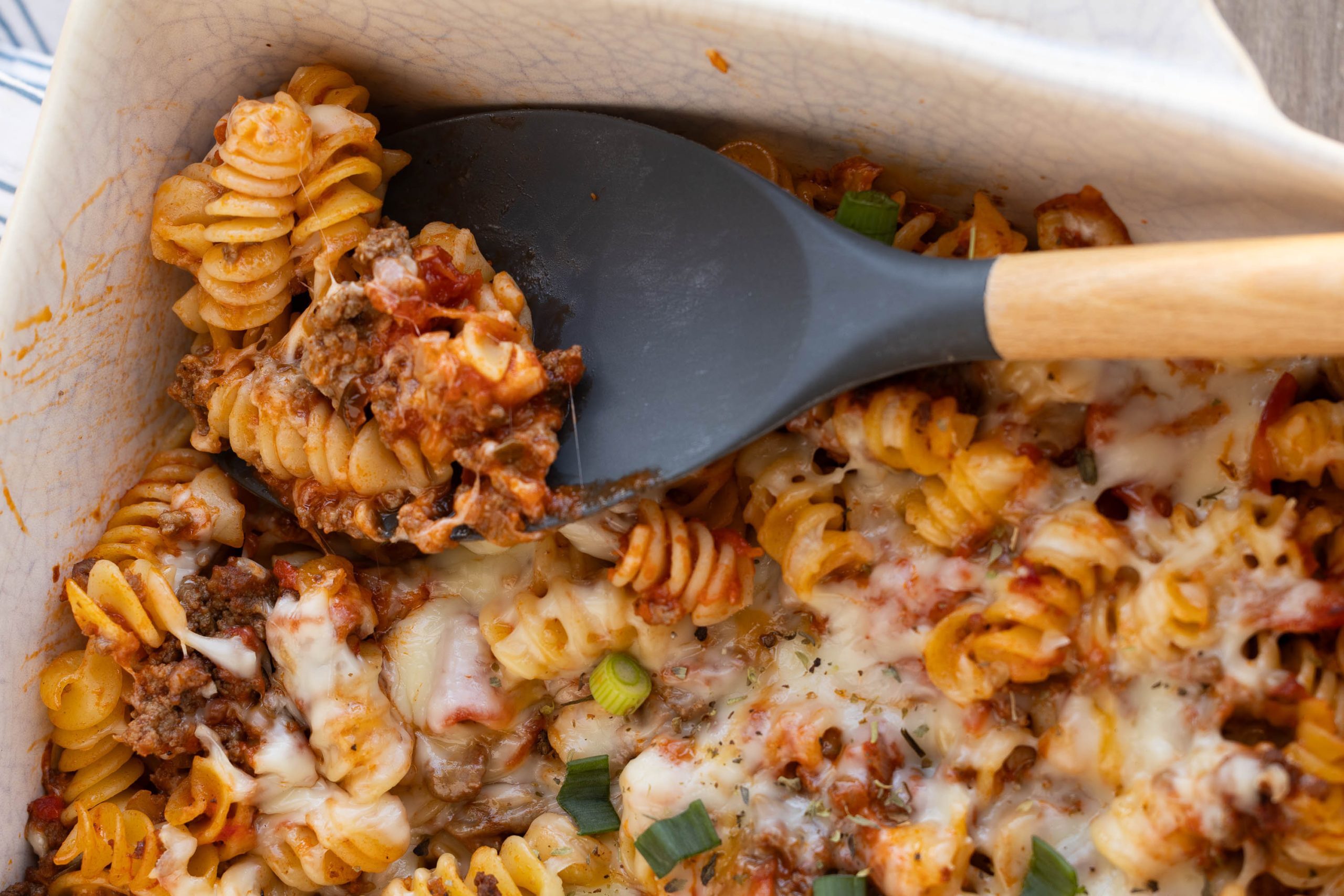 A close-up of baked rotini pasta with melted cheese, ground meat, tomato sauce, and green onions in a casserole dish, with a serving spoon scooping a portion.