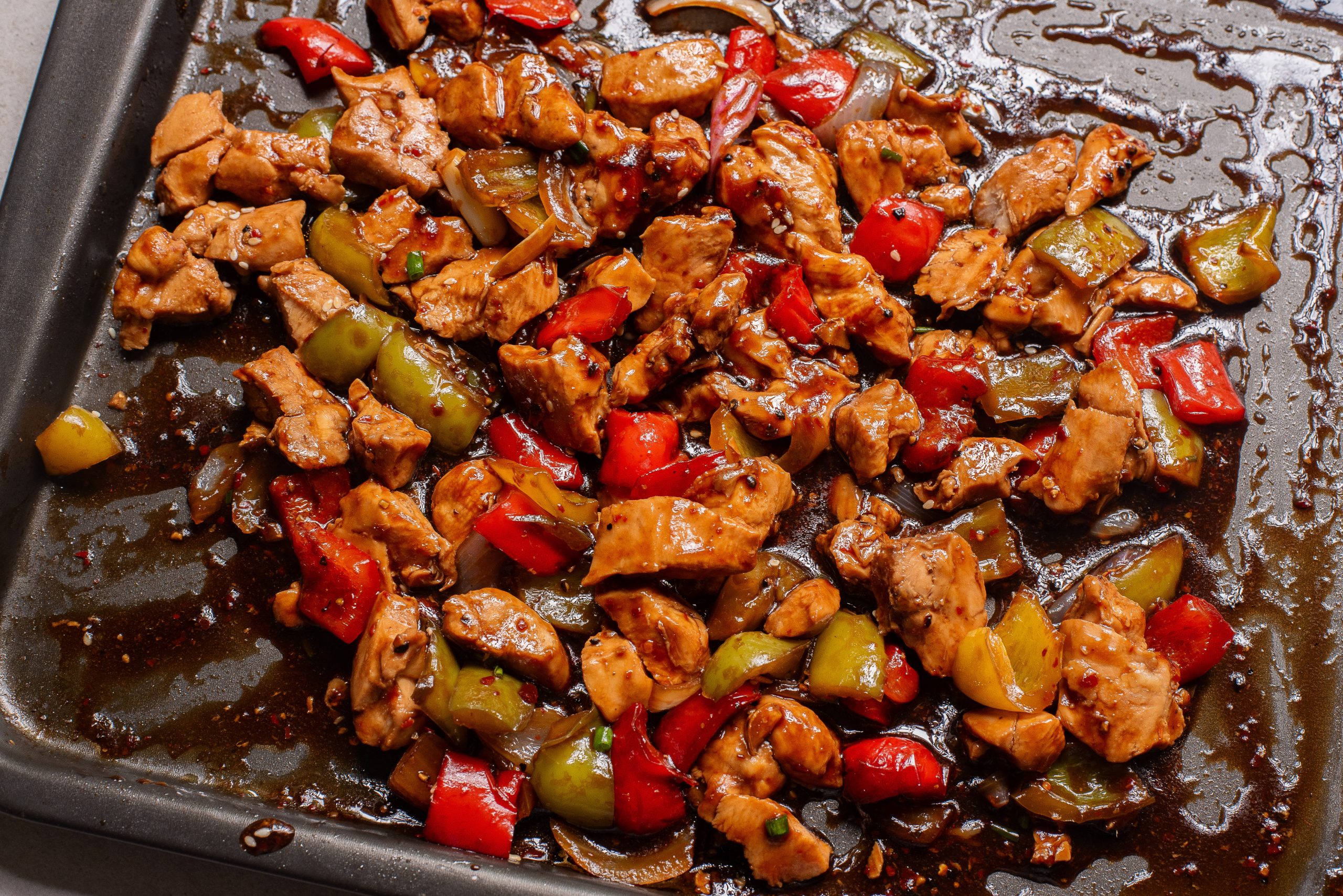 Sheet Pan Bell Pepper Chicken features chopped cooked chicken with red and green bell peppers in a dark sauce on a baking sheet.
