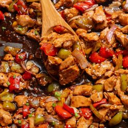 A wooden spoon scoops a savory mix reminiscent of Sheet Pan Bell Pepper Chicken—diced chicken, red and green bell peppers, and onions coated in a dark sauce—from a pan.