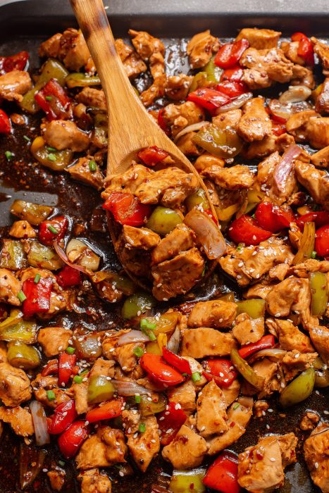 A wooden spoon scoops a savory mix reminiscent of Sheet Pan Bell Pepper Chicken—diced chicken, red and green bell peppers, and onions coated in a dark sauce—from a pan.