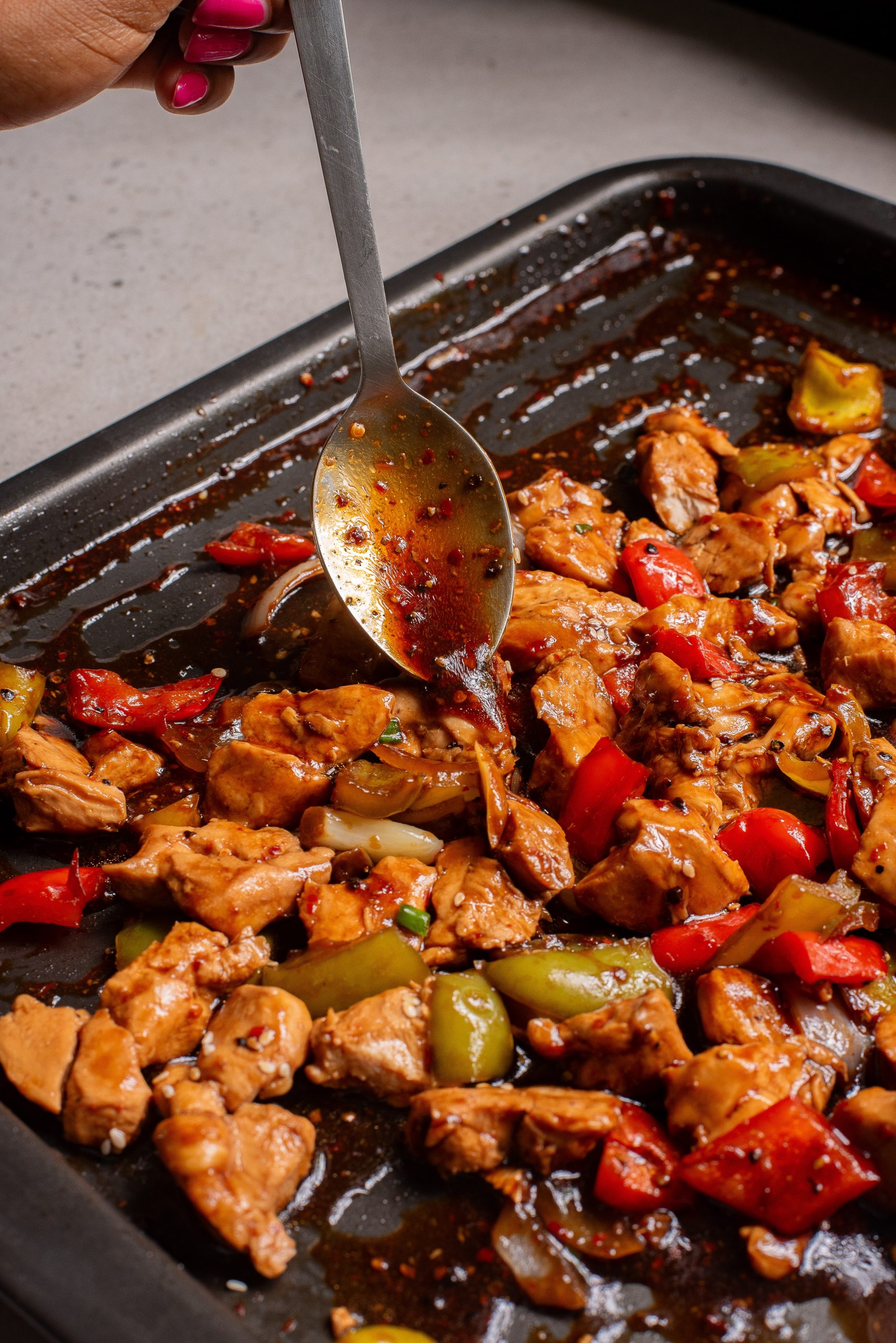 A hand holds a spoon over a tray with a savory sauce.