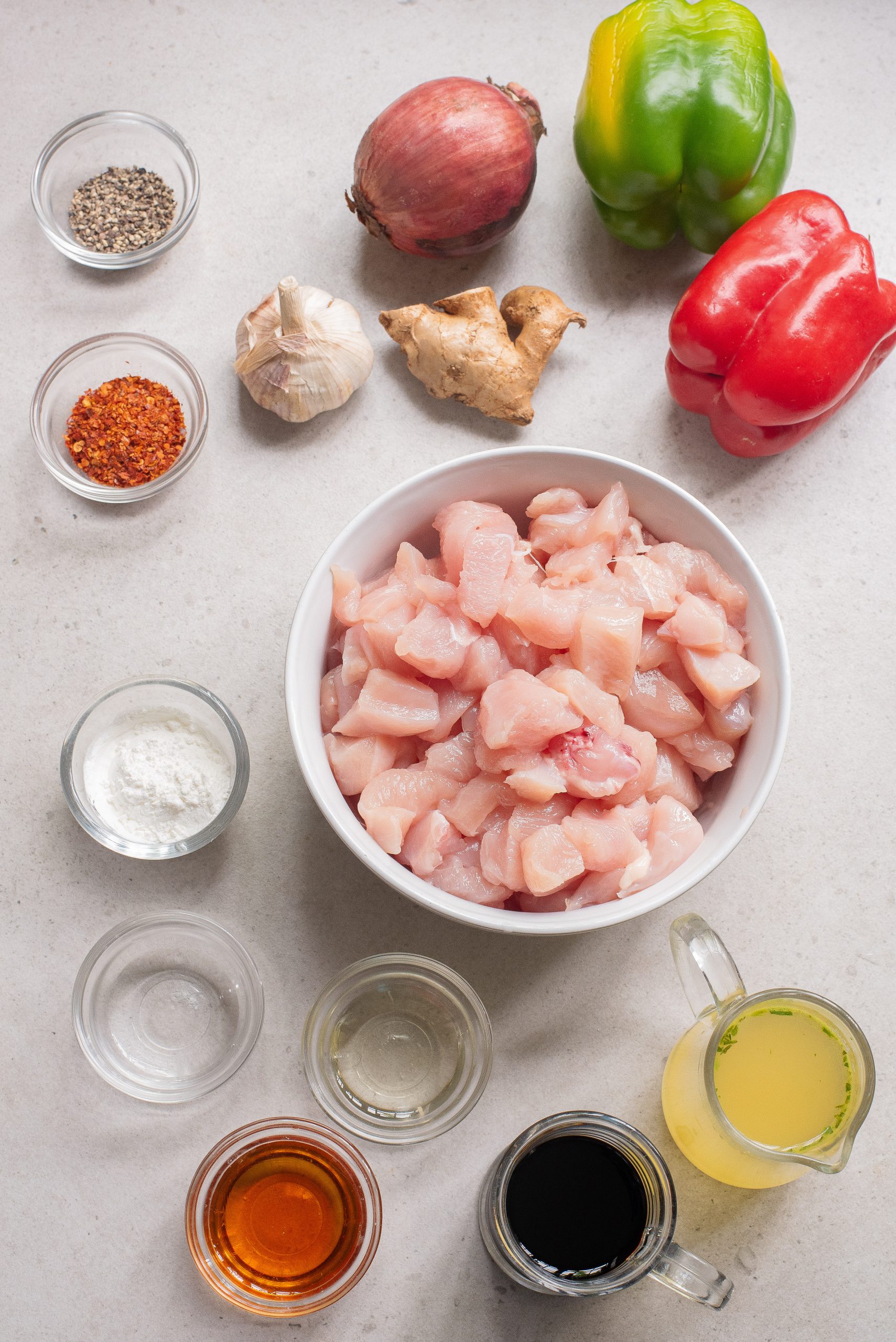A bowl of diced raw chicken, perfect for Sheet Pan Bell Pepper Chicken, is surrounded by spices, liquids, garlic, ginger, a shallot, and vibrant green and red bell peppers on a countertop.